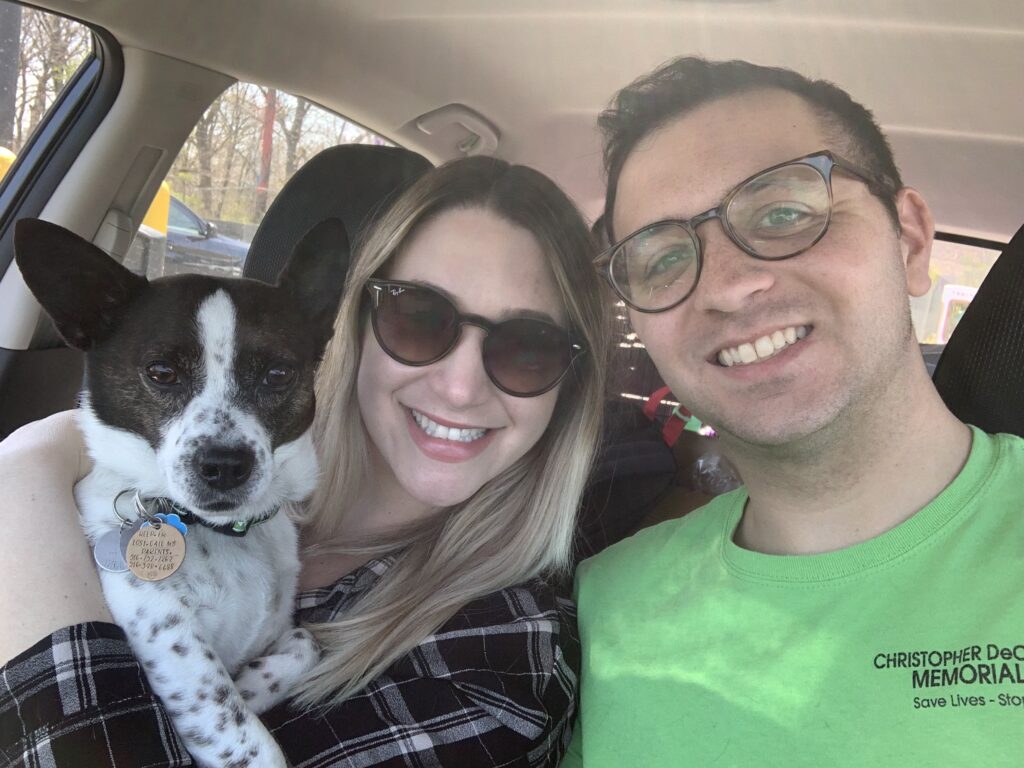 A man in a green shirt and glasses, and a woman in a flannel shirt with sunglasses smile for a selfie in a car. The woman holds a black & white spotted dog.