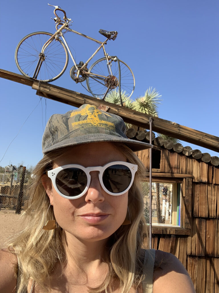 A woman with blonde hair, round white sunglasses, and a grey hat poses outdoors, under a sculpture with a bicycle.