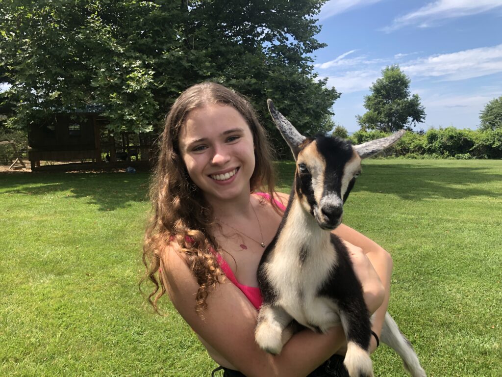 Ari - A Young woman with wavy brown hair smiles and holds a baby goat.