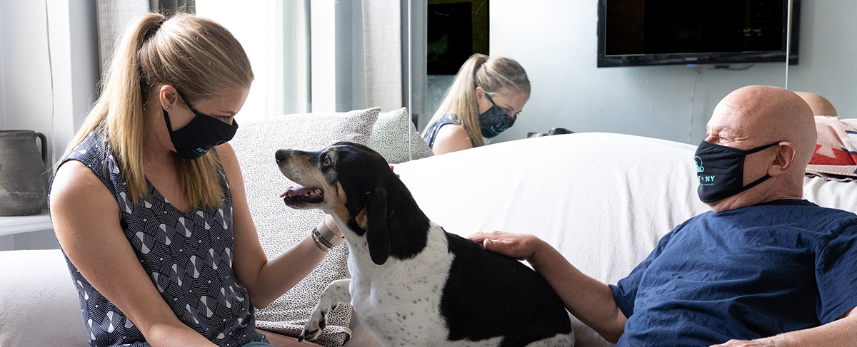 A young blonde woman in a grey patterned shirt sits on the couch with an older bald gentleman in a blue t-shirt, with a Brown & Black Basset Hound / Beagle mix dog in the middle. Both are wearing face masks and looking at the dog, who is looking at the woman.