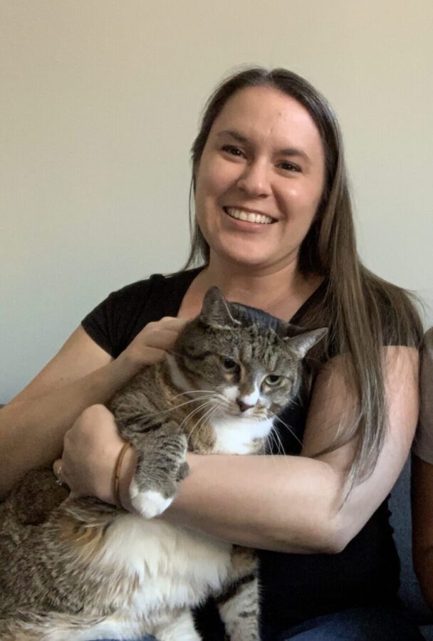 Kimberly, a woman with long dark hair and brown eyes, smiles at the camera while holding her cat Franklin, a white, brown, and grey tabby cat who looks toward the ground. Kimberly is the Community Outreach Manager for PAWS NY.