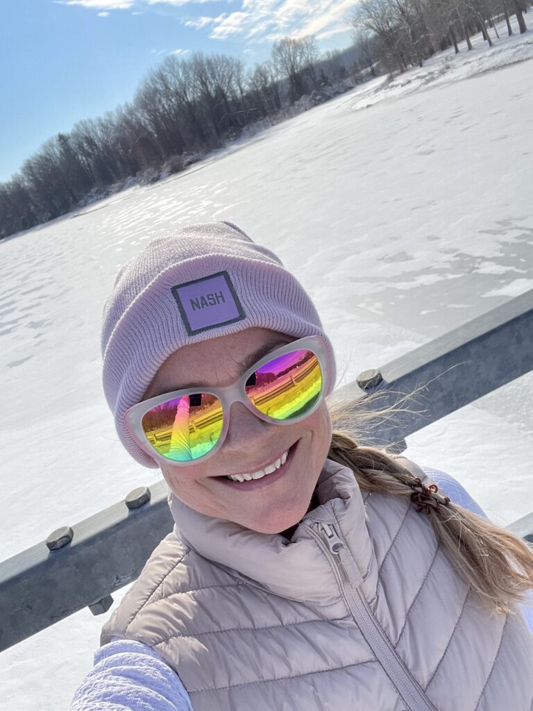 A woman in a light pink puffy vest and hat, with rainbow sunglasses, takes a selfie and smiles at the camera, with a field covered in snow behind her.