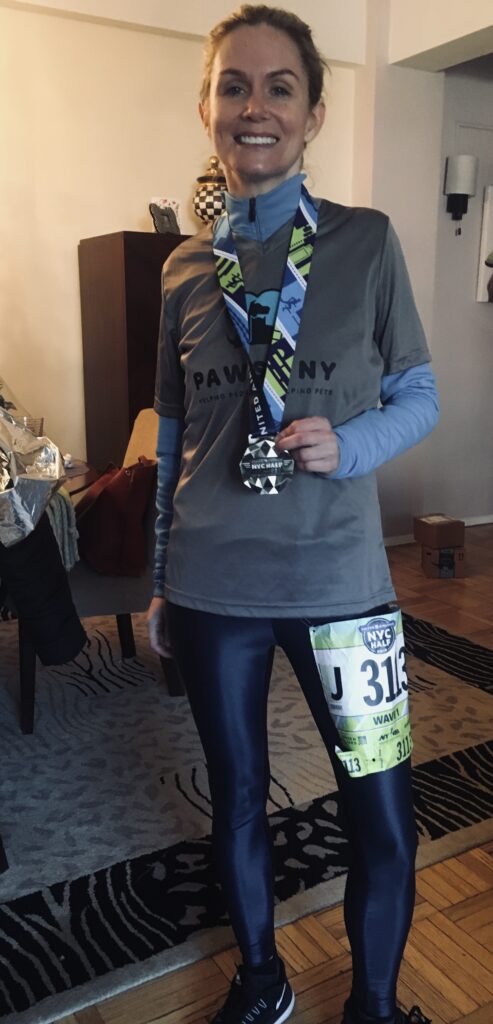 A woman (Amy) in running gear and with her hair back, stands posing with a race medal from the NYC Half. She's wearing a grey shirt with the PAWS NY logo.