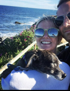 With cliffs and the ocean in the background, a woman with reflective sunglasses smiles at the camera, holding a dog that is looking off to the right. A man squeezes into the frame. 