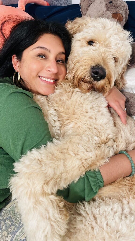 A young woman with dark hair, hoop earrings, and a green sweater hugs her large Goldendoodle dog. They both smile at the camera.