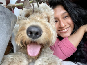 A large dog, a Goldendoodle, fills the frame with his face, with a black nose and pink tongue sticking out. A young woman in a pink shirt is seen in the background, smiling and snuggling with the dog.