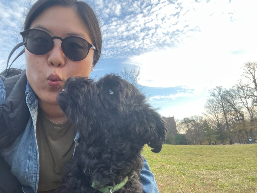 Connie Volunteer Spotlight - A woman with dark hair and sunglasses makes a kissy face at the camera, while a dog with black curly fur leans up to kiss her face. They are outside in a grassy field with a lightly cloudy sky.