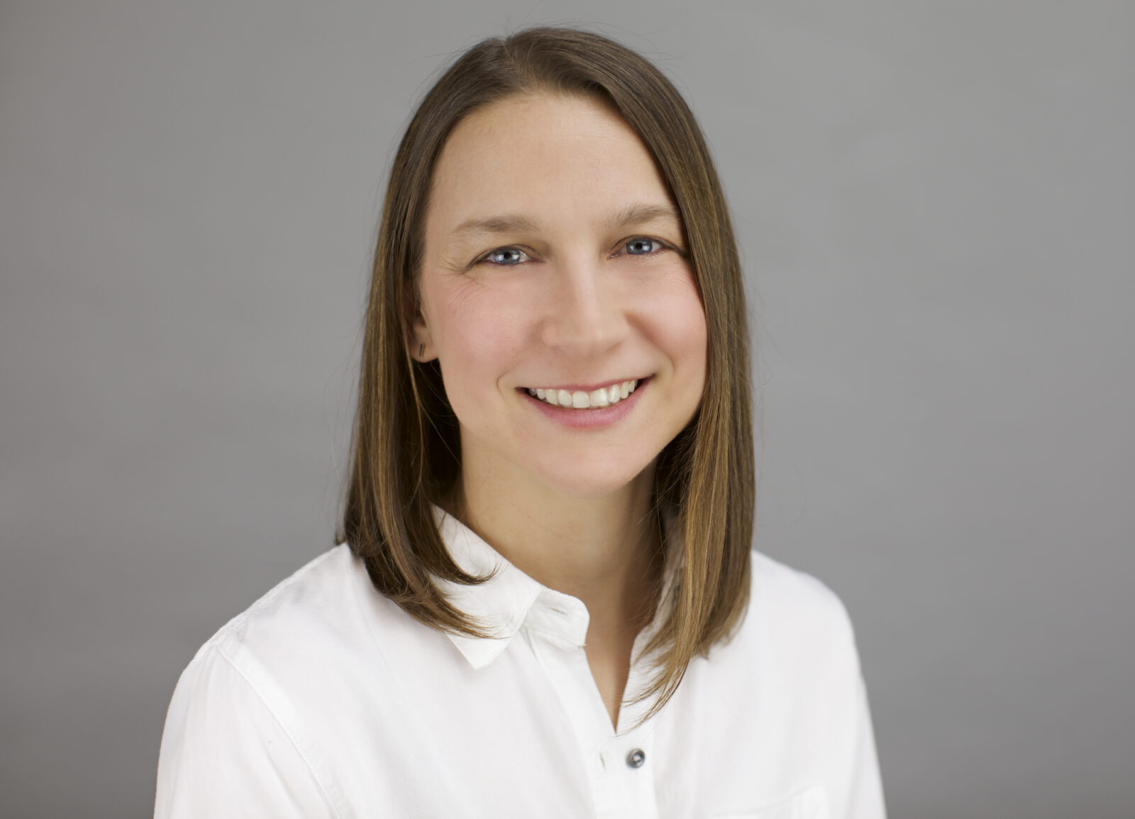Rachel Herman's headshot by Katie Kinsley - A woman with straight shoulder-length brown hair smiles at the camera in a white button down shirt. The picture is from her mid-torso up.