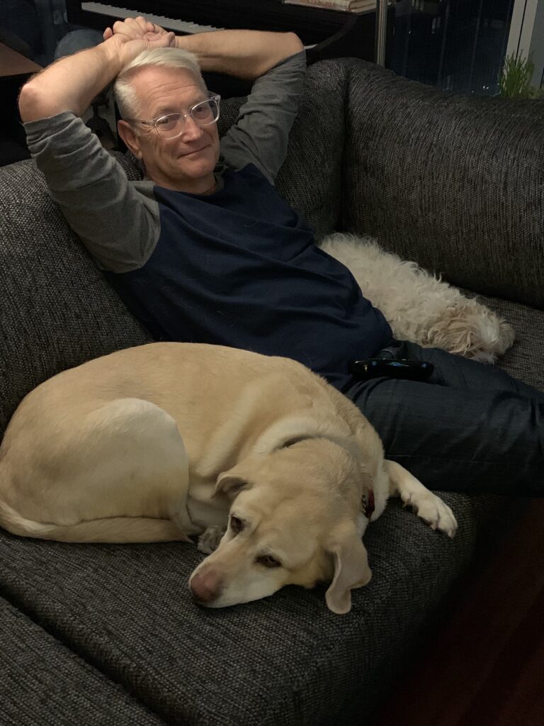 Scott Volunteer Spotlight - A man with silver hair and glasses in a dark long-sleeved shirt lounges and lays back on a couch, with a small dog tucked up on his left side, and a larger yellow lab curled up on his right side.