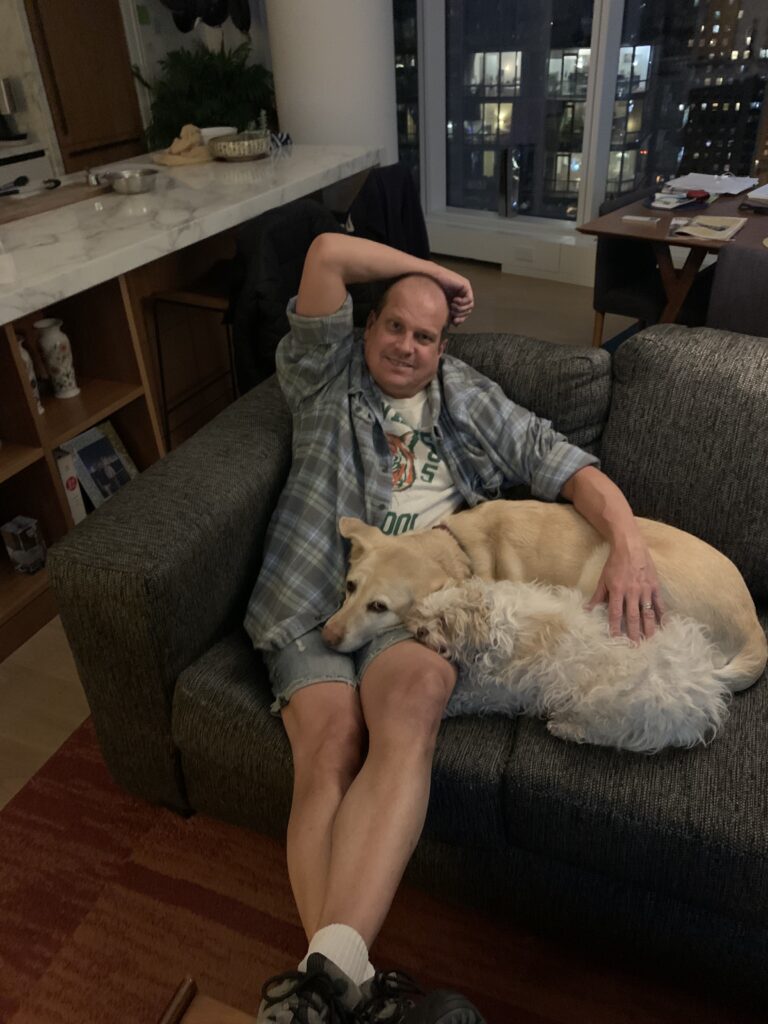 Scott's Husband Ed - A man in a patterned button down shirt and shorts sits on a couch with two dogs laying their heads in his lap: a small white dog and a bigger yellow lab. 
