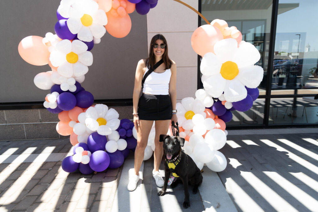 A woman with dark hair and sunglasses stands in front of a balloon archway with her dog, a black lab mix. She is wearing a white tank top and black shorts, while the dog has a yellow harness.