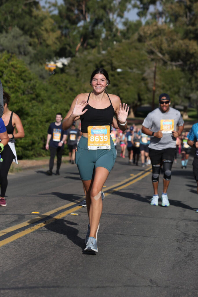 Shay, a tall woman with dark hair, wear a black top and green shoes and shorts as she runs in a race in a park.