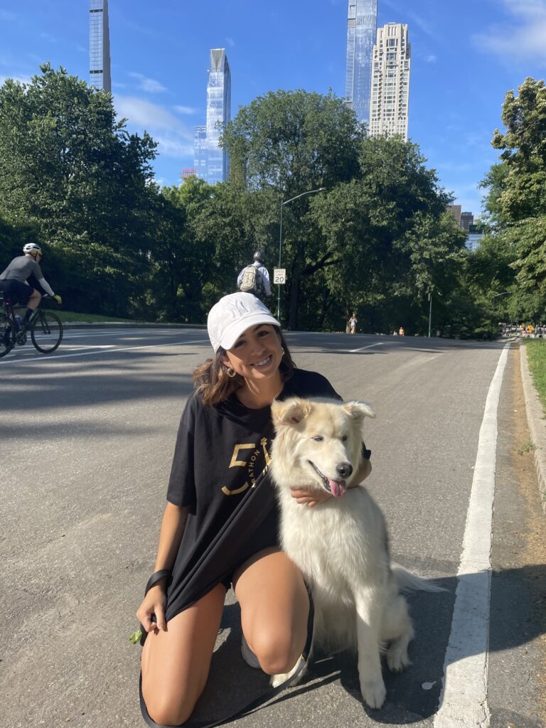 A young woman in a black 50th TCS NYC Marathon shirt and a white hat crouches in the road with her arm around a white husky/samoyed mix dog. Both are smiling at the camera, with Central Park and NYC buildings in the background.