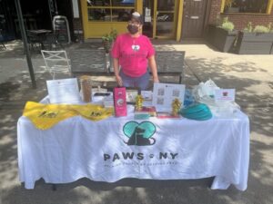A woman in a bright pink PAWS NY t-shirt wears a black hat and face mask and stands behind a table with a PAWS NY tablecloth and other giveaways on it. She's outdoors, at a street fair.