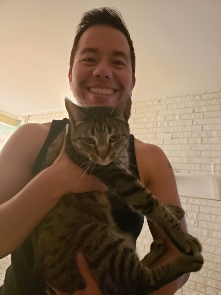 A smiling man with dark hair looks into the camera and holds up a brown tabby cat.