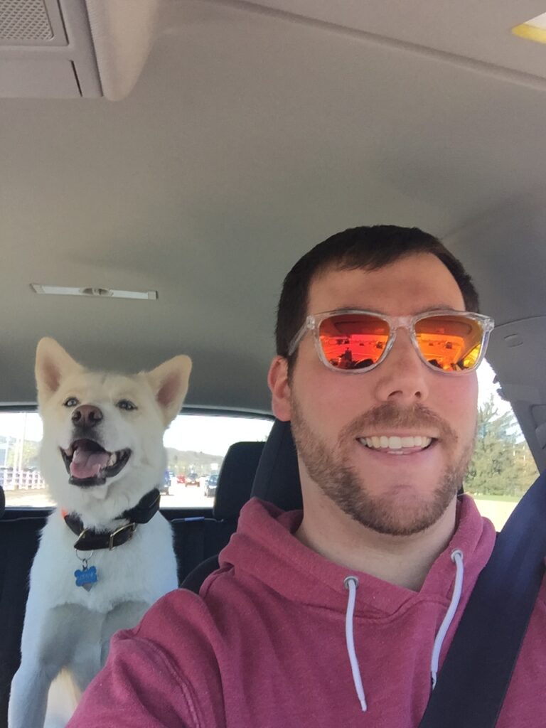 Michael, a young man in a red hoodie with orange reflective sunglasses, sits in a car, with a white dog in the seat behind him.