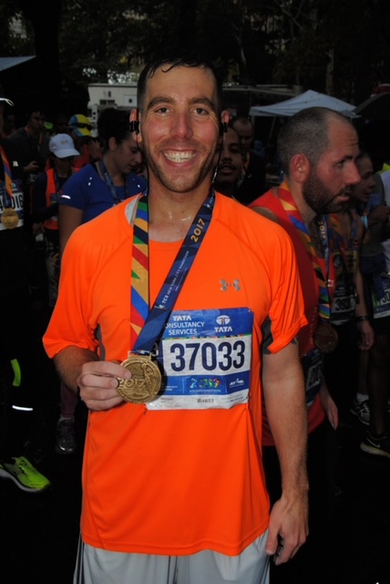 Michael, a tall and slim smiling young man, wears a bright orange t-shirt and holds a medal from a race. He just finished the race and is surrounded by other runners.