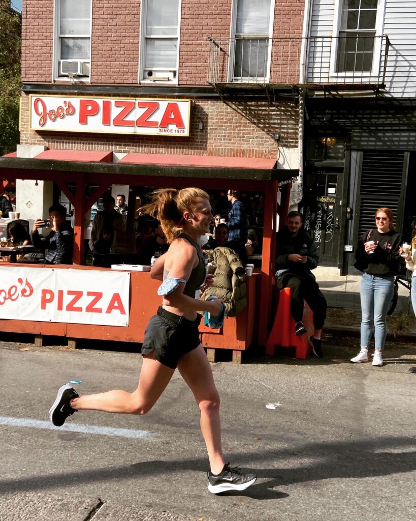 Annie runs on a street in New York City, wearing black shorts and running shoes, and a black sports bra. Her red hair is up in a messy ponytail / bun. She runs past a Joe's Pizza restaurant, with people out in front cheering her on.