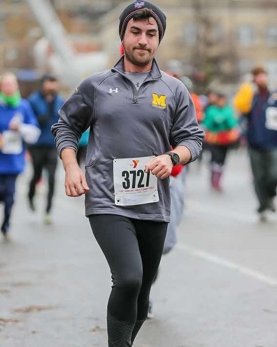 Justin, a young man in a grey University of Michigan long-sleeved shirt and dark grey hat, runs a race outdoors. He has black pants and a white runner's bib with the numbers 3727.