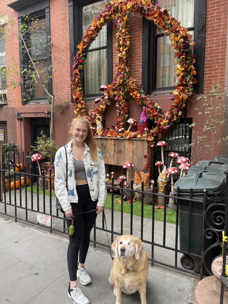 The outside of a New York City apartment is decorated with a fall theme: red and white polka dot mushrooms; orange, red, and green leaves and flowers in the shape of a large peace sign. Annie, who has long red hair pulled back into a ponytail, stands in front of the display, with Golden Retriever Valla on a leash beside her. Annie is wearing black leggings and running shoes, with a grey cropped top and a light blue jean jacket. 
