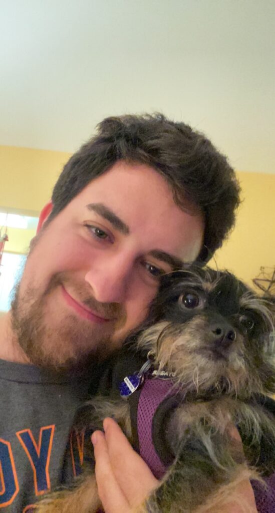Justin, a young man with dark brown hair, brown eyes, and a light brown beard, takes a selfie with a dog. He tilts his head to the right and smiles closed-mouthed at the camera. He leans his head against a scruffy brown and black dog that is wearing a purple harness and looking off to the right.