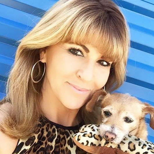 Gigi, a woman with blonde / light brown hair with bangs and loose waves, looks at the camera with a close-mouthed smile. She wears hoop earrings and a leopard print top, with a blue background. She holds a small dog, who has a leopard-print toy in its mouth.