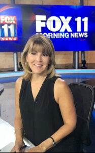 Gigi sits at a News Desk, with signs for FOX 11 Morning News in the background. She wears a black v-neck top, with thin bracelets and hoops earrings. Her hair is a medium-length layered cut with bangs and is blonde / light brown.