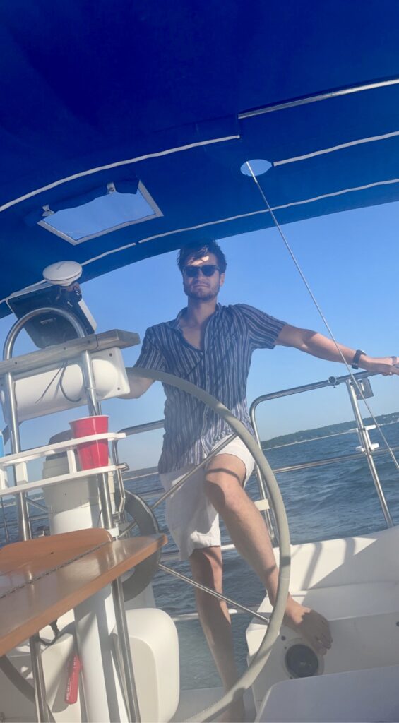 Harrison is a young man with dark hair and sunglasses. He is wearing a vertical striped shirt and white shorts, while at the helm of a sailboat. He has one hand on the helm and leans the other on a railing of the boat.
