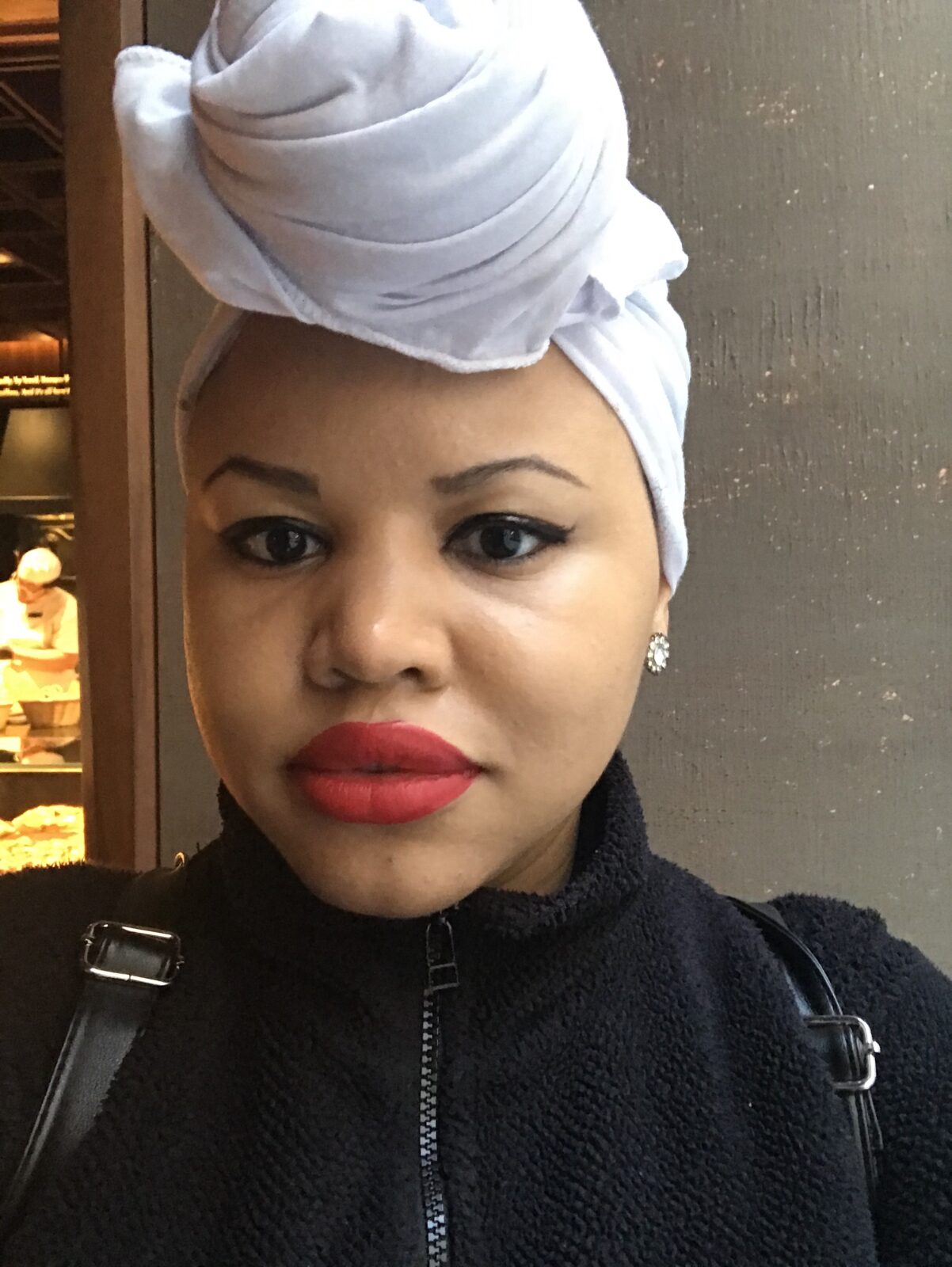 A selfie of Simone, a young black woman wearing a black zip-up sweatshirt and backpack straps. She has on red lipstick and her hair is in a white hair wrap, knotted to the front.
