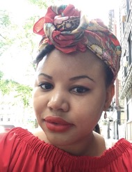 A close-up selfie of Simone, a young black woman wearing a patterned headscarf tied in the front and red lipstick that matches her top. She's standing on a sidewalk with the city in the background.