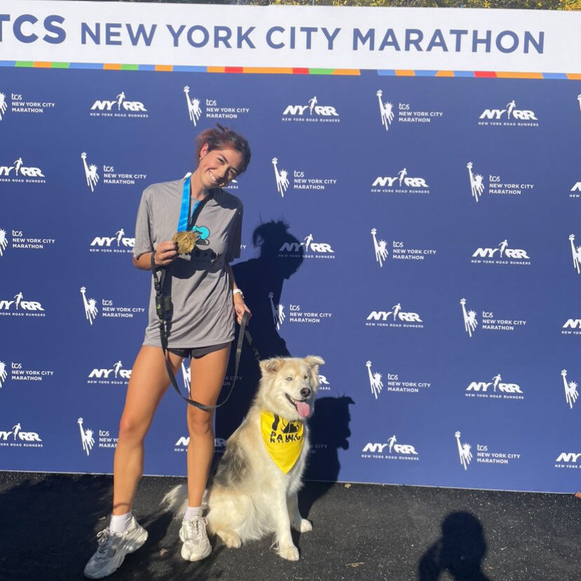 A young woman wearing a grey PAWS NY shirt and running shorts / shoes stands in front of a TCS New York City Marathon step & repeat, showing off her medal and posing with her dog, who is wearing a yellow PAWS NY bandana.