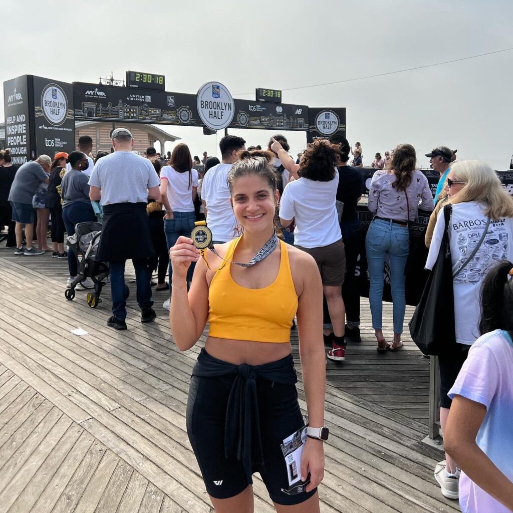 A woman in a yellow tank top and black shorts poses on the Coney Island Boardwalk at the end of the Brooklyn Half Marathon, holding up her finisher's medal.