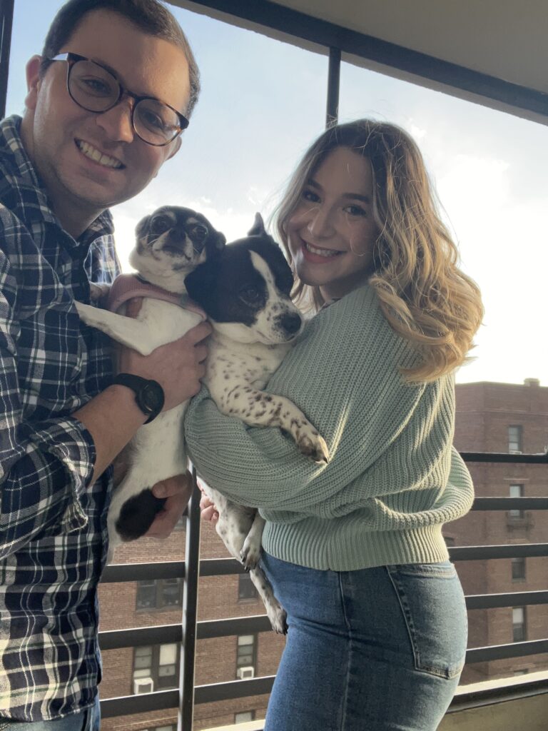 Kyle and his wife stand next to a window holding their two dogs. 
