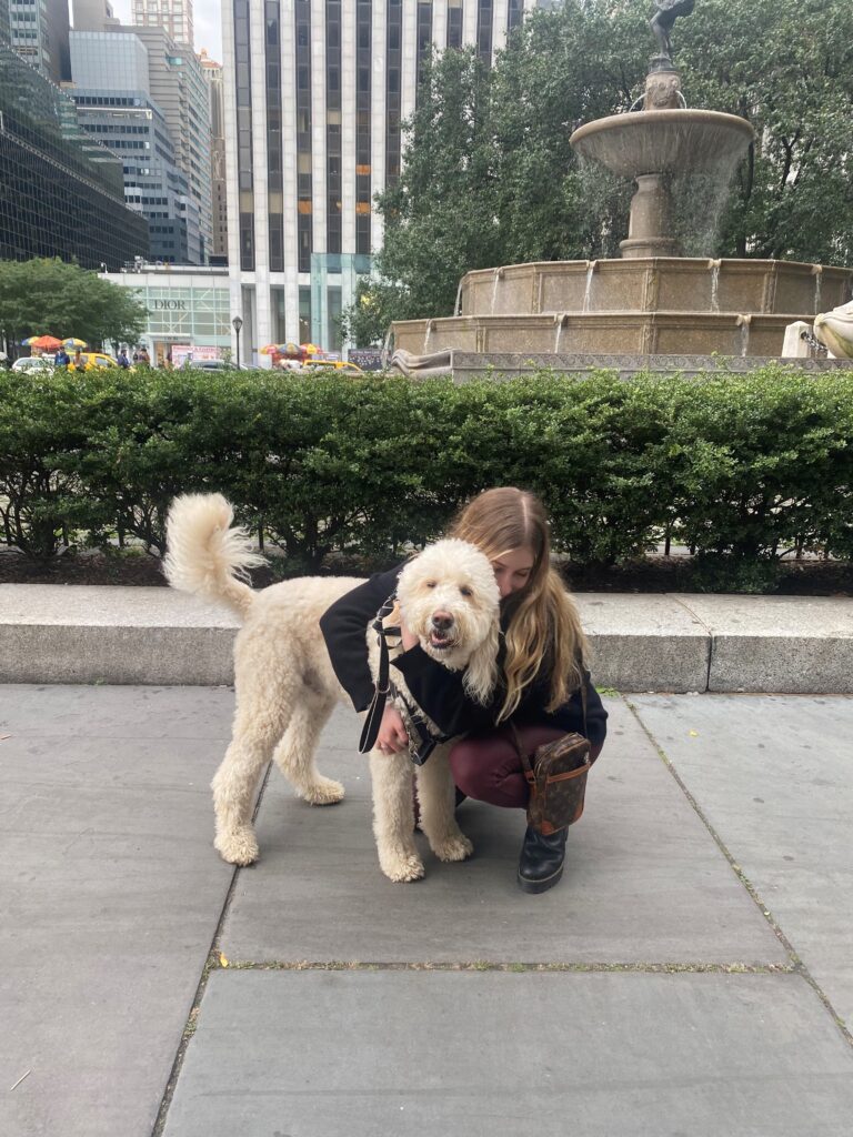 Cody and Saree - a young woman with long light brown hair crouches on the sidewalk in front of a fountain. She hugs a light colored goldendoodle, who stands on all fours and looks at the camera.