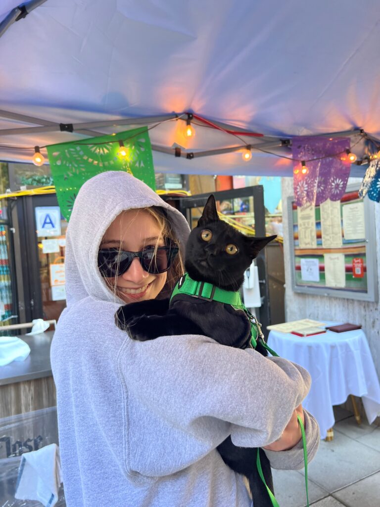 Onny and Saree - A young woman in a gray hoodie and black sunglasses holds up a black cat in a green harness.