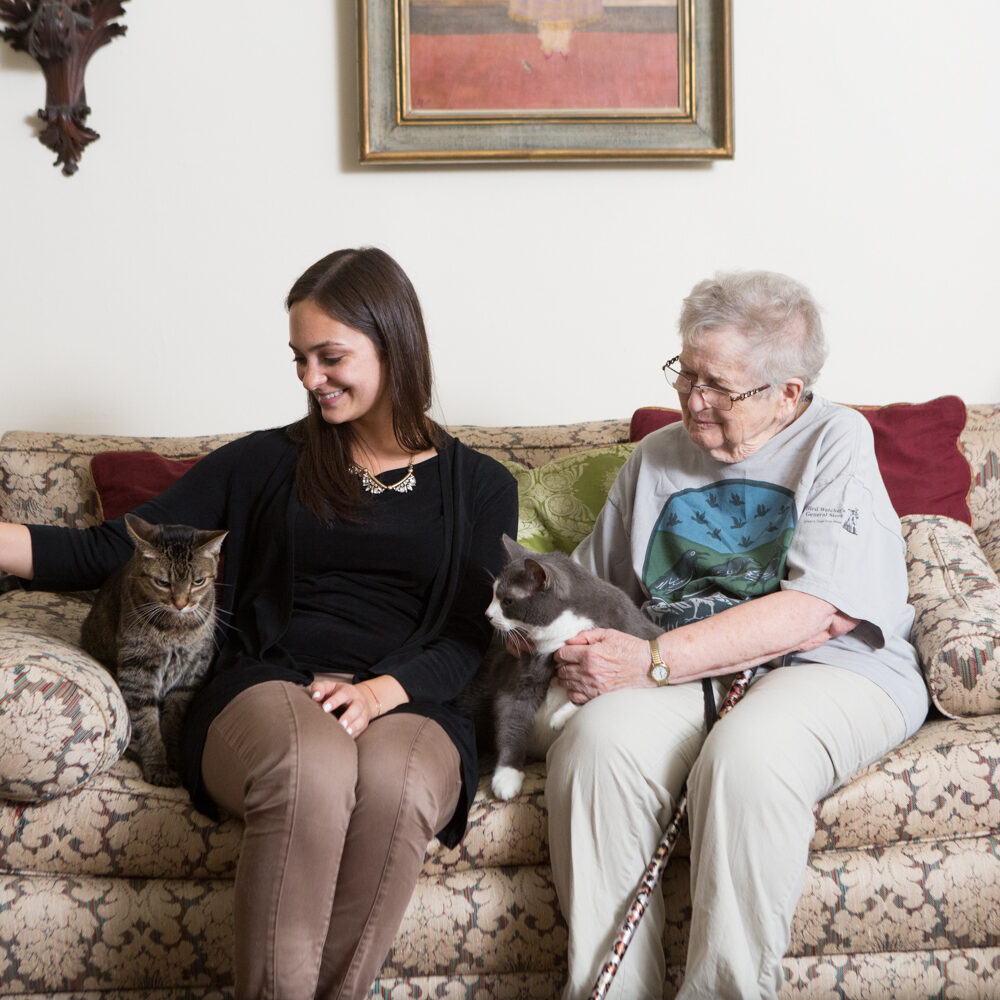 A PAWS NY volunteer with dark hair, a black long-sleeved top and brown pants, sits on a floral couch with her arm around a brown and grey tabby cat. An older woman with white hair and glasses sits next to her, holding a grey and white cat.