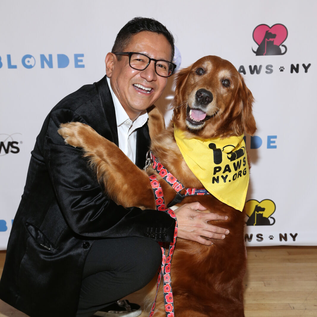 Cesar Fernandez-Chavez and Louboutina the Hugging Dog attend PAWS NY 2019 Spring Benefit. Cesar is wearing a suit and is crouched down next to Louboutina, who is wearing a floral harness and leash, with a PAWS NY bandana around her neck. Both are smiling at the camera and posed in front of a logo-ed backdrop.