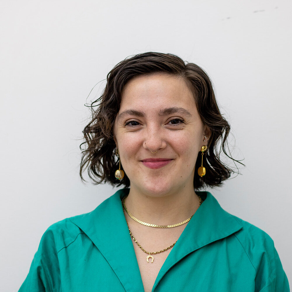 Headshot for Elia Rangel - A woman with curly / wavy brown hair that is chin length wears a teal button down shirt with gold earrings and several necklaces.