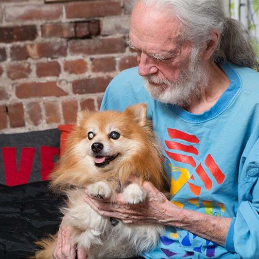 PAWS NY About Us - An older gentleman with long grey/white hair and a beard, holds his dog, a pomeranian mix , while the dog smiles with its tongue out.